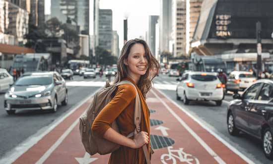 Utlandsstuderande tjej som står med en skolryggsäck i mitten av ett övergångsställe i en storstad. Runt om syns bilar och höga byggnader
