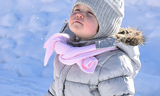 Litet barn som står med grå mössa och jacka och rosa halsduk som vänder huvudet mot den fallande snön