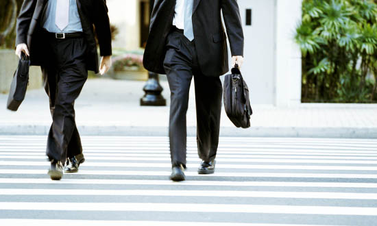 Benen på två affärsmän som går på ett övergångsställe. De håller i varsin portfölj.