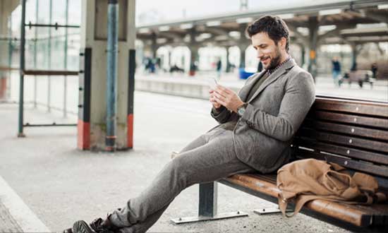 Man i kostym som sitter på bänk på tågstation och väntar. Han kollar i sin mobiltelefon. 