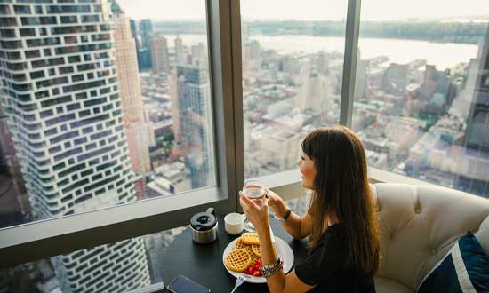 Kvinna som sitter på hotellrum och kollar ut genom ett stort fönster med en kopp kaffe i handen