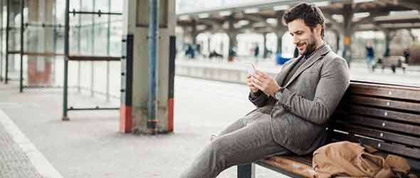 Man i kostym på tågstation. Han sitter på en bänk och tittar på sin mobiltelefon