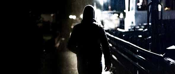 Mörk bild på en man i luvtröja bakifrån. Han går i regnet. 