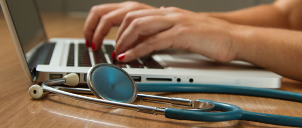 Händer som syns skriva på en laptop. I förgrunden ligger ett stetoskop.