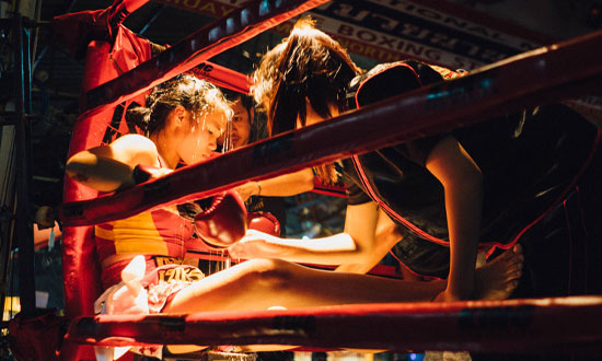 Kvinnlig boxare som sitter och blundar i ringhörnan. Två personer ser över henne.