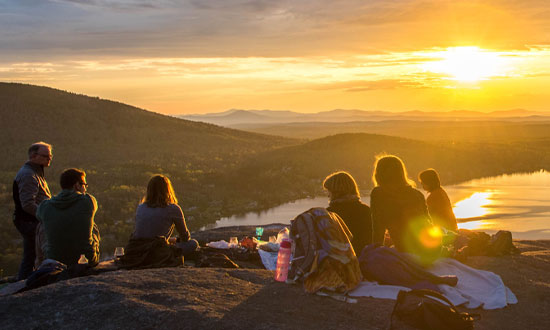Backpackervänner som sitter och äter på en höjd och ser ut över en dal i solnedgång.